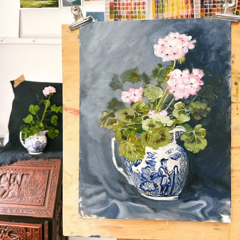 A still life painting of pink geraniums in a blue and white patterned vase, displayed on an easel in a studio setting.