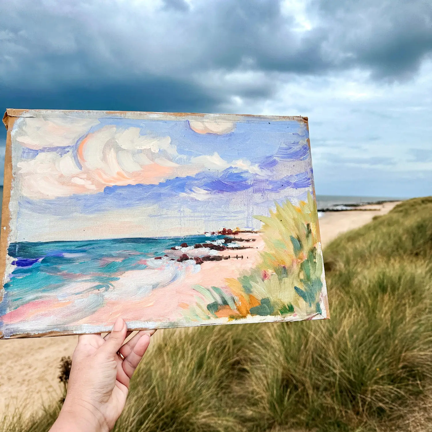 A hand holds a small oil painting of a beach and sea under a cloudy sky, with a real beach dune landscape blurred in the background. Painted en plein air in Norfolk, UK.