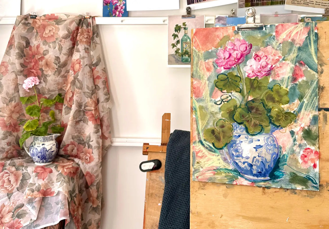 A textured loose mixed media painting of pink flowers in a blue and white vase, displayed on an easel next to a floral-patterned chair in an artist's studio. Still life studio set up example.