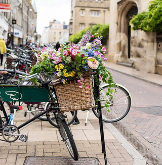 A black vintage-style bicycle with a green cargo box and a large woven basket overflowing with colorful flowers, parked on a city sidewalk next to a stone street.