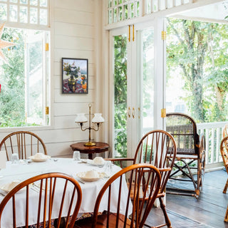 Queenslander interior photo with a still life painting by Rebecca Eddison on the wall. A bright dining room corner with a white tablecloth, wooden spindle-back chairs, a small wooden side table with two white lamp shades, and large windows and glass doors opening to a lush green outdoor porch.